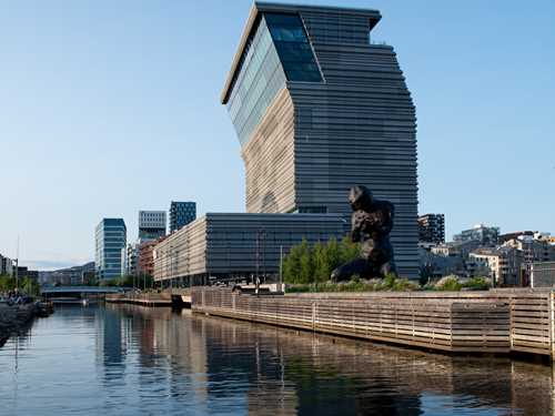 MUNCH-bygget sett fra sørsiden i strålende solskinn. Bygget speiler seg i vannet og du ser skulpturen The Mother i forkant av bildet. Fotografi