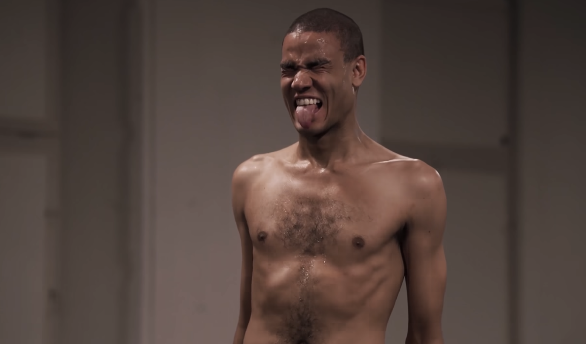 Harald Beharie under fremførelsen av en performance. Han er i halvfigur og bar overkropp. Kroppen er spent og han kniper øynene sammen og rekker tungen ut i en grimase. Fotografi