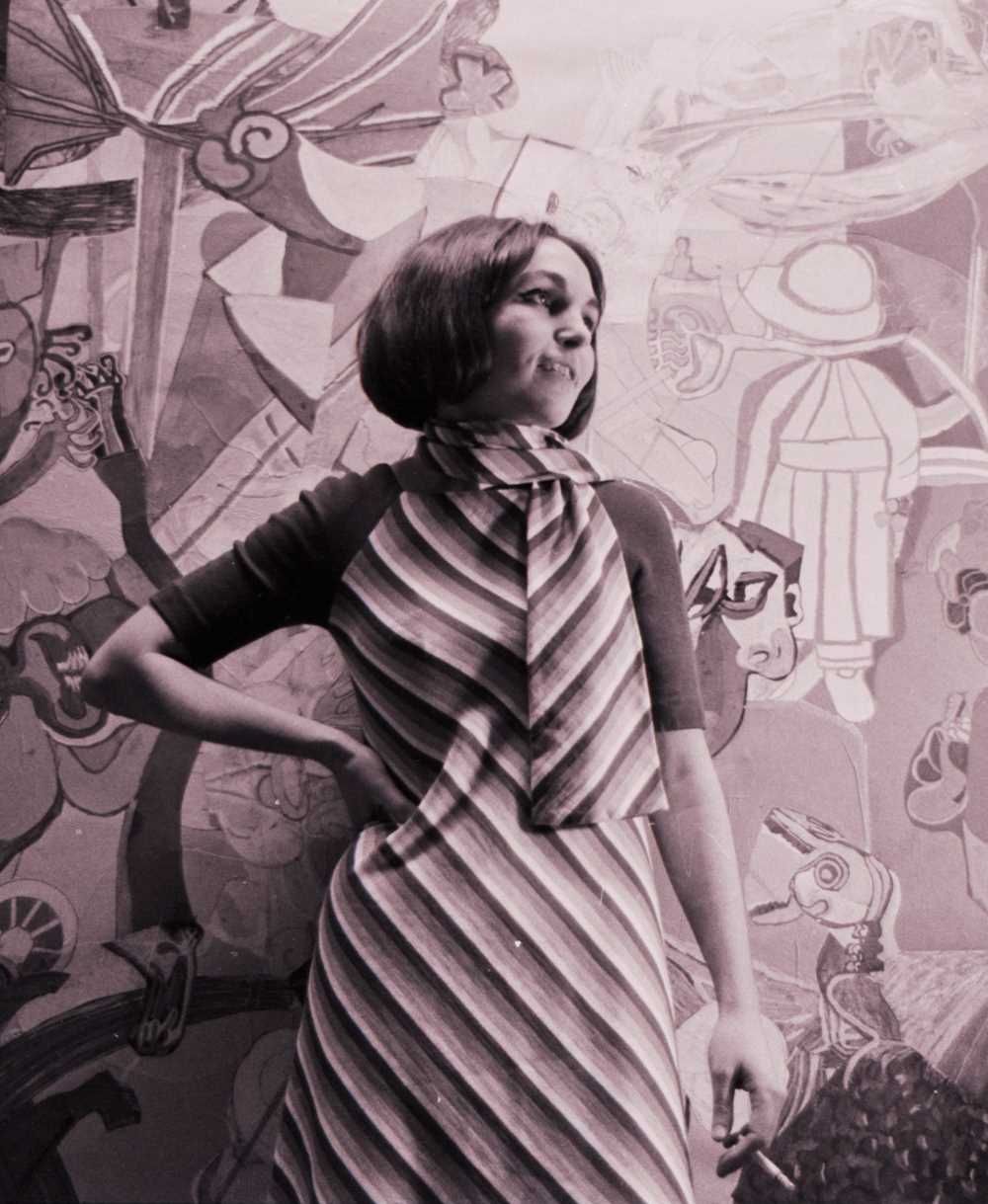 Black and white photograph of a young woman (Paula Rego). She is wearing a striped dress and has her right hand on her hip while looking to the side.