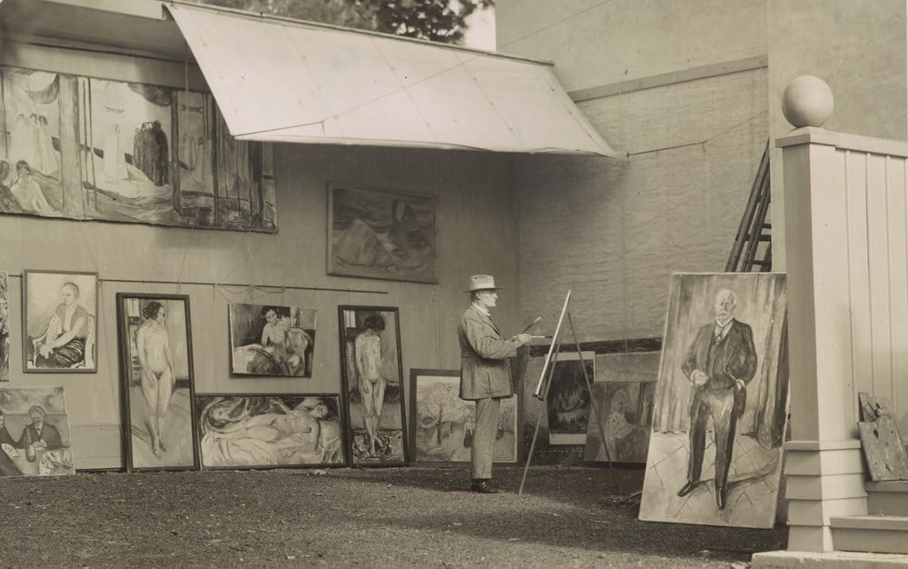 Edvard Munch is standing and painting in his open-air studio at Ekely. He is surrounded by his own works and is wearing a hat to shield himself from the sun and a light-coloured suit.