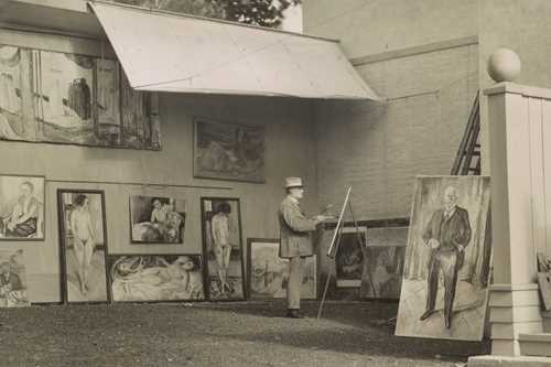 Edvard Munch is standing and painting in his open-air studio at Ekely. He is surrounded by his own works and is wearing a hat to shield himself from the sun and a light-coloured suit.