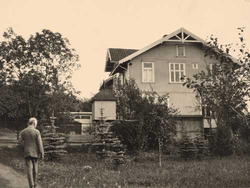 Edvard Munch vandrer langs veien oppover mot Ekely. Det er en gresseng og trær foran huset.