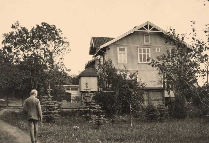 Edvard Munch walks along the road leading up to Ekely. There is a grassy meadow and trees in front of the house.