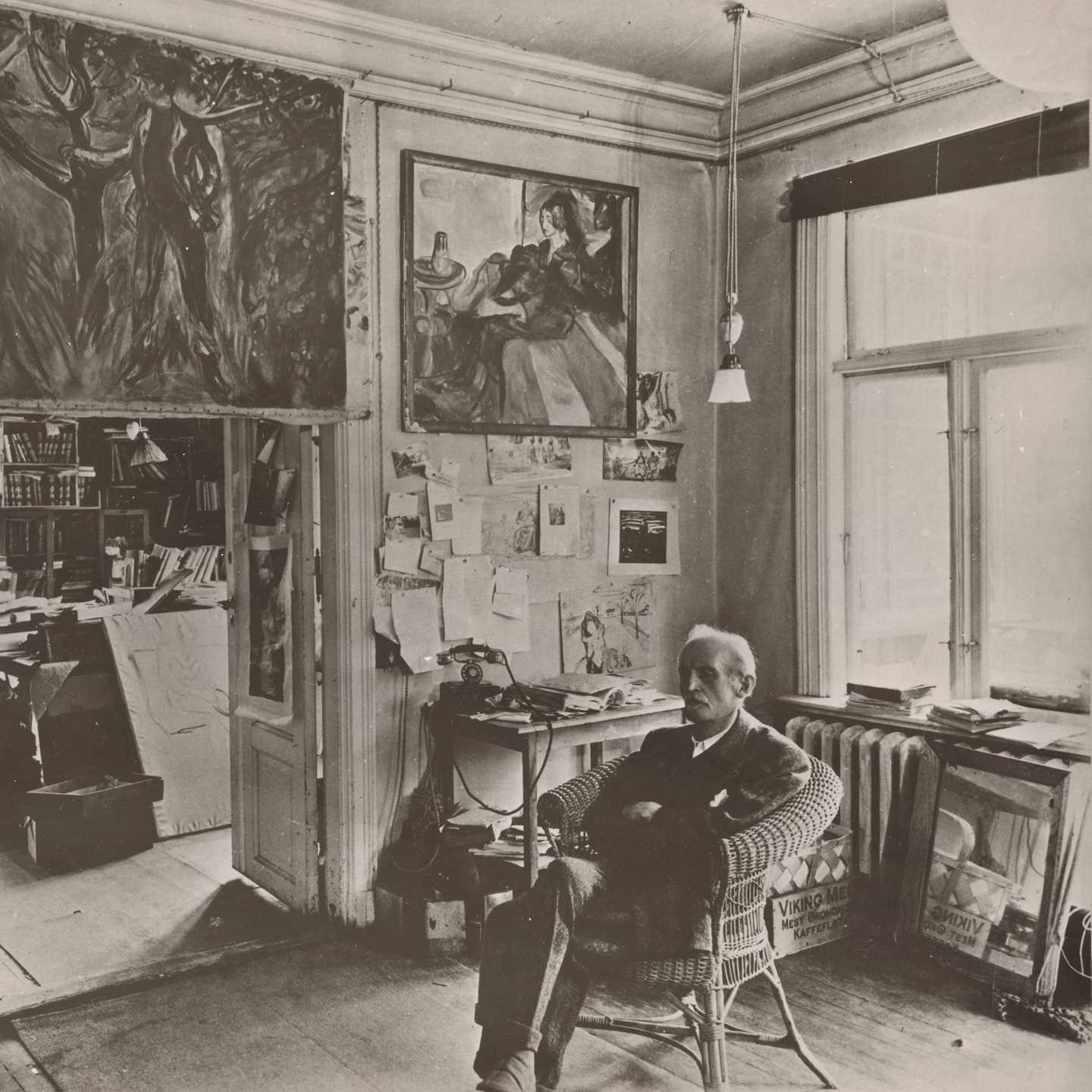 Edvard Munch sits in a wicker chair in a room at Ekely, surrounded by his paintings. Photograph.
