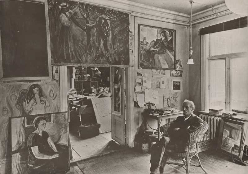Edvard Munch sitter i en kurvstol i et rom på Ekely omgitt av sine malerier. Fotografi