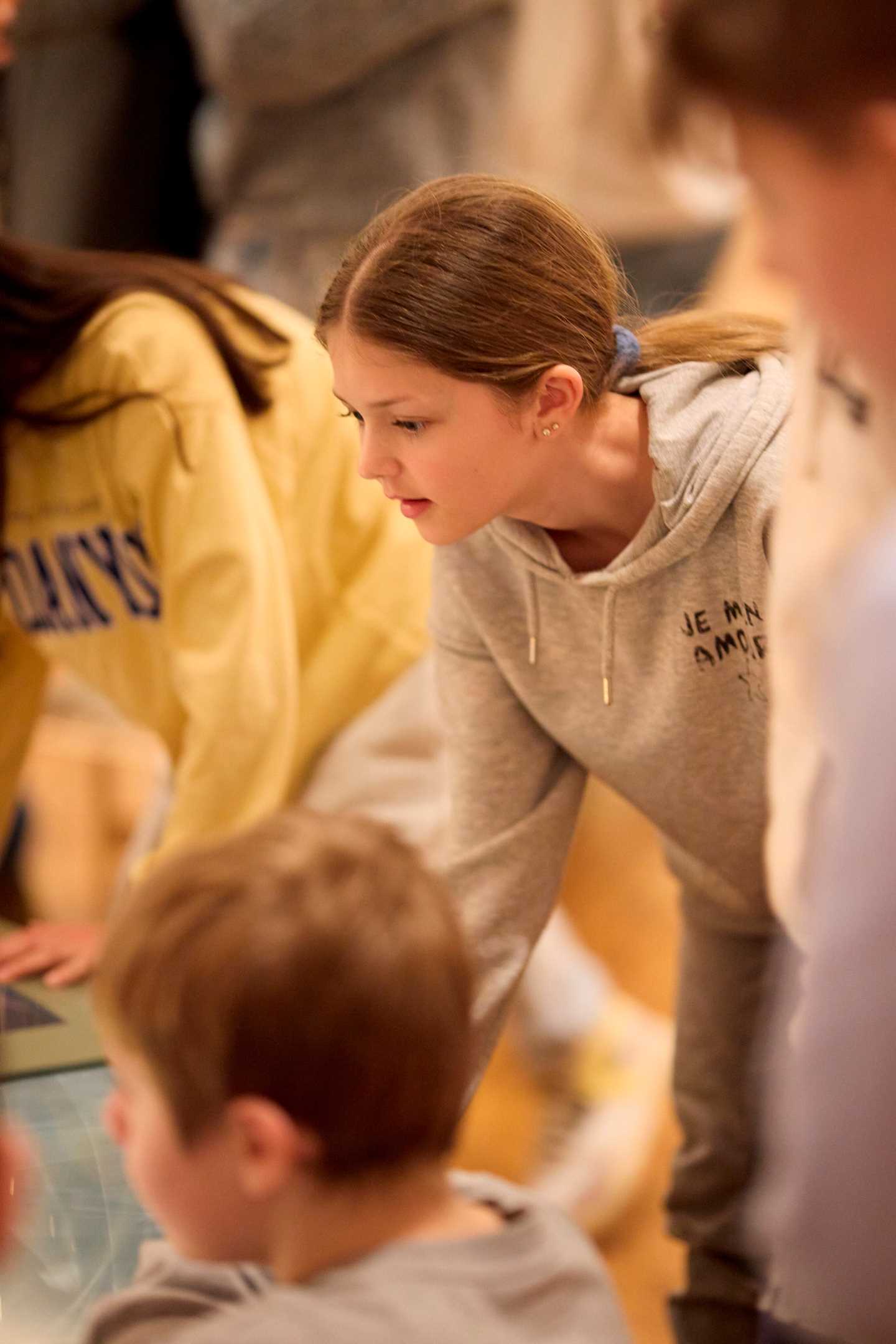En ung jente ser interessert på et bord med en glassplate, rundt henne er det flere ungdommer. Fotografi.