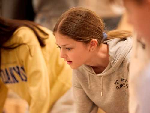En ung jente ser interessert på et bord med en glassplate, rundt henne er det flere ungdommer. Fotografi.