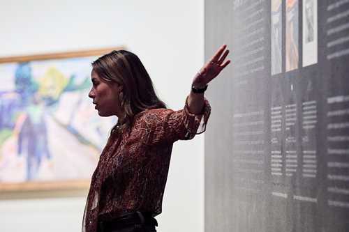 A mediator at MUNCH stands in front of a blue wall with text explaining the different versions of The Scream held by the Munch Museum. Photograph.