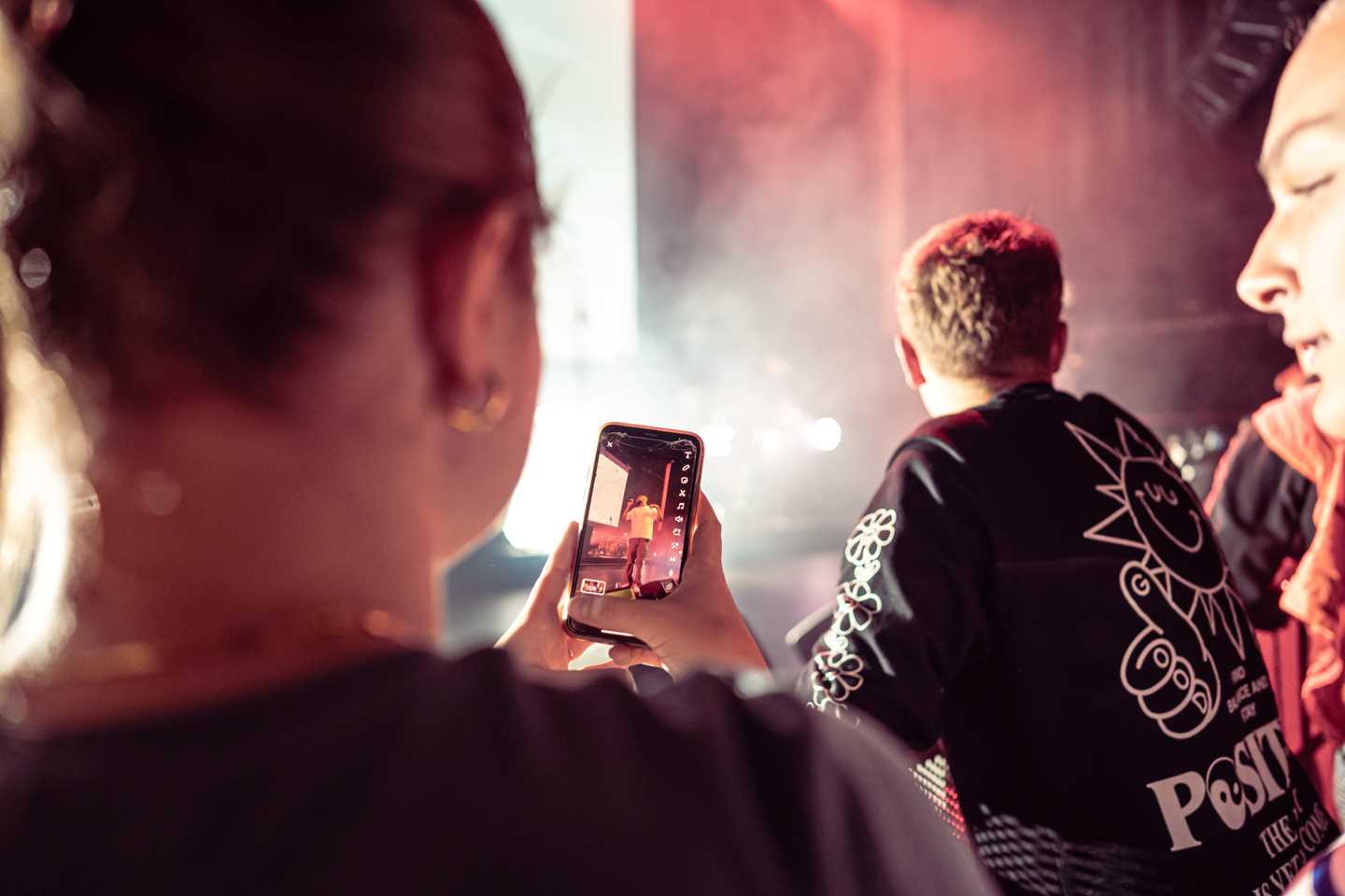 Unge mennesker på konsert. En tar bilde av artisten på scenen med mobilen. Fotografi