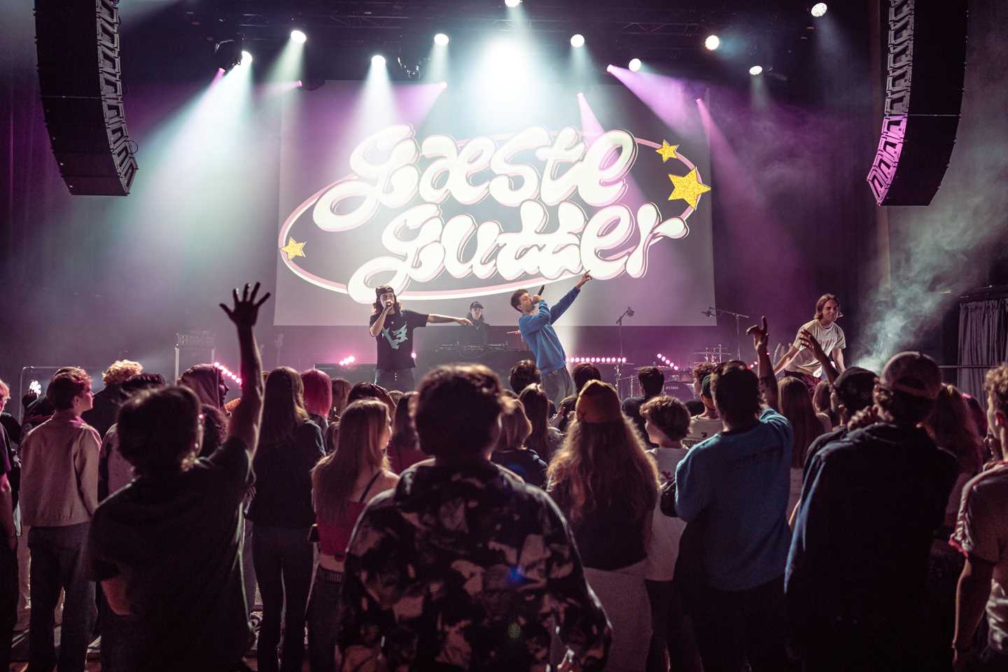 En full konsertsal med mennesker som står med ryggen mot kamera og ser på en scene der flere hiphop-artister opptrer. Det er lilla scenelys og bak scenen henger et backdrop der det står "Gæste gutter". Fotografi