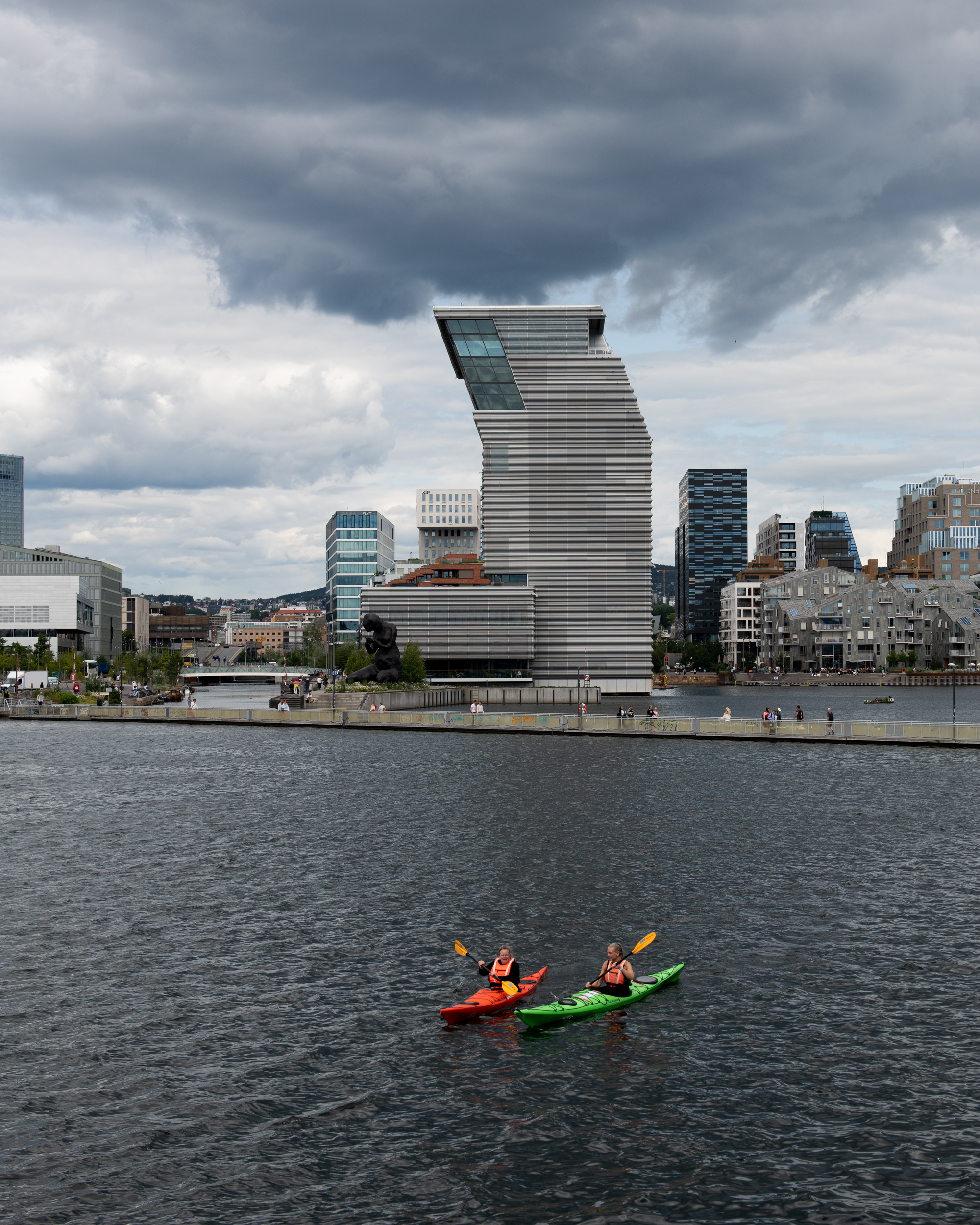 MUNCH seen from Sørenga. In the foreground, two people paddle kayaks, with a floating walkway behind them. The sky is blue-grey and cloudy. Photograph.
