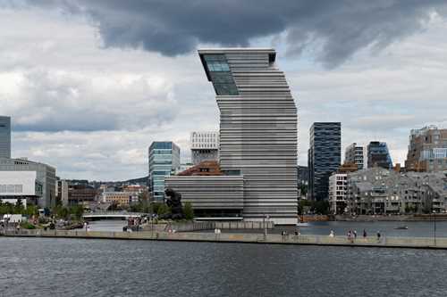 MUNCH seen from Sørenga. In the foreground, two people paddle kayaks, with a floating walkway behind them. The sky is blue-grey and cloudy. Photograph.