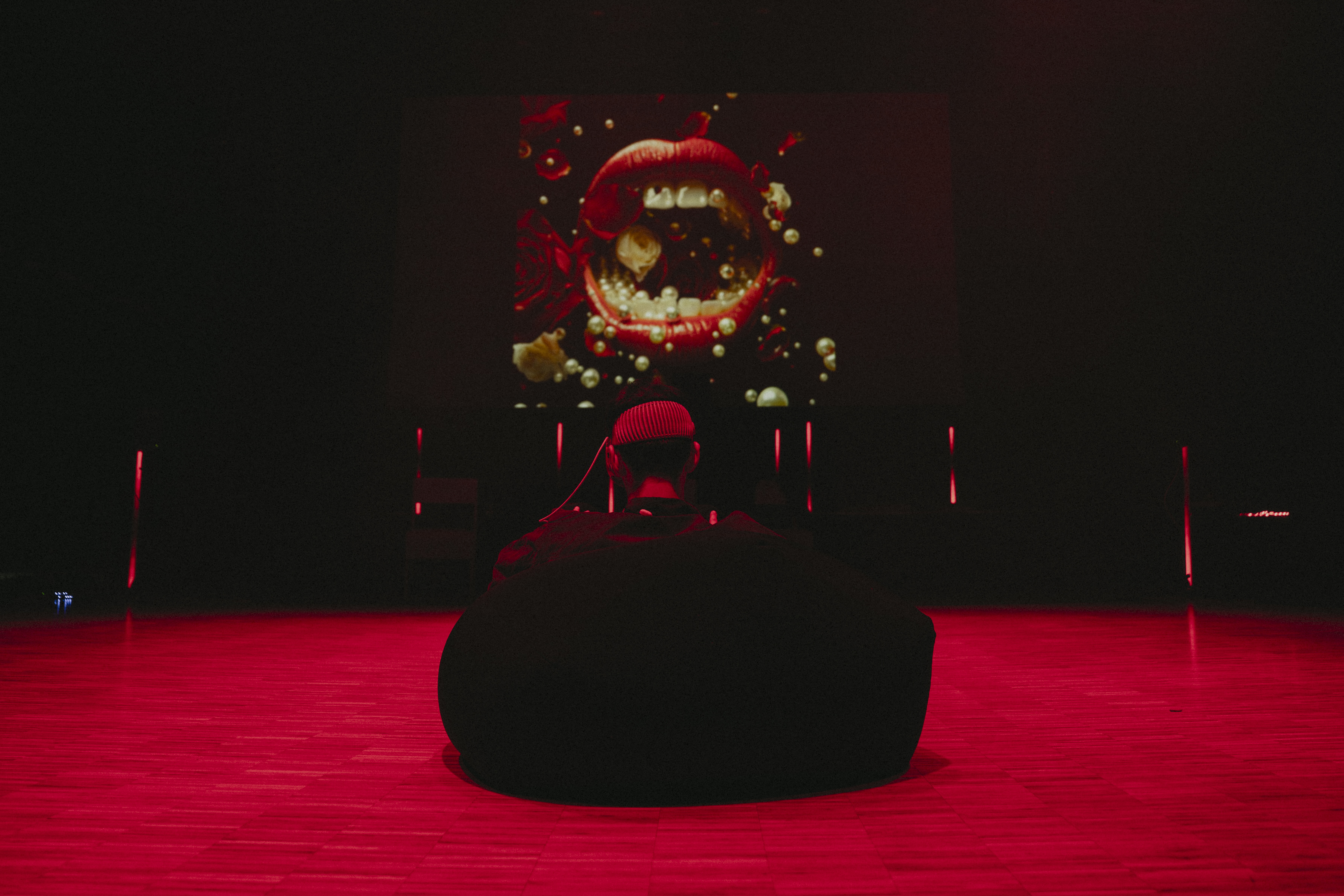 En person sitter med ryggen mot kamera i en sakkosekk på gulvet. Rommet er lyssatt så gulvet ser rødt ut. På veggen fremfor personen er det et bilde som viser en stor, åpen, munn med røde lepper og hvite tenner. Fotografi