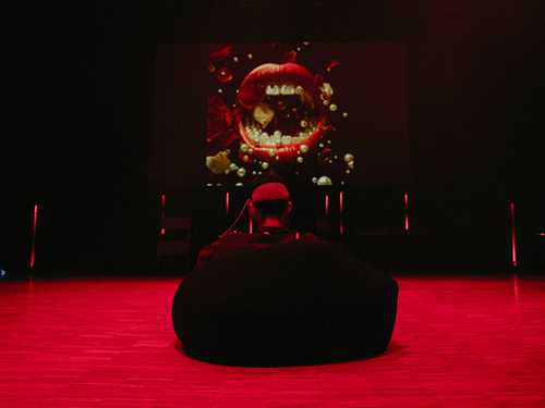 A person sits with their back to the camera in a beanbag chair on the floor. The room is lit so that the floor appears red. On the wall in front of the person is an image of a large, open mouth with red lips and white teeth. Photograph.