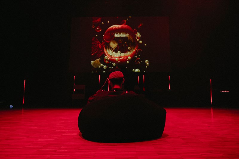 En person sitter med ryggen mot kamera i en sakkosekk på gulvet. Rommet er lyssatt så gulvet ser rødt ut. På veggen fremfor personen er det et bilde som viser en stor, åpen, munn med røde lepper og hvite tenner. Fotografi