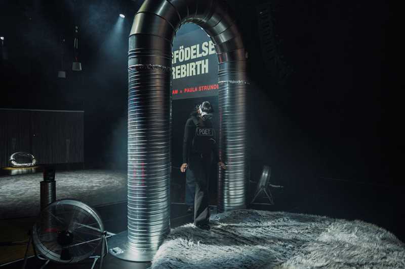 A woman moves through a portal made of ventilation pipes while wearing VR goggles. Photograph.