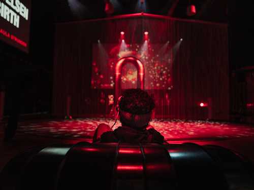 A boy sits with his back to the camera wearing VR goggles. He is looking towards a portal illuminated in red light. Photograph.
