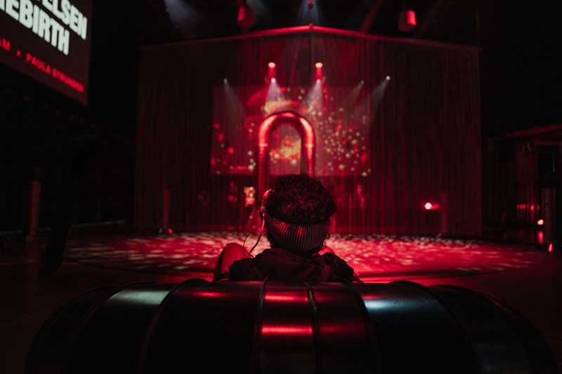 A boy sits with his back to the camera wearing VR goggles. He is looking towards a portal illuminated in red light. Photograph.