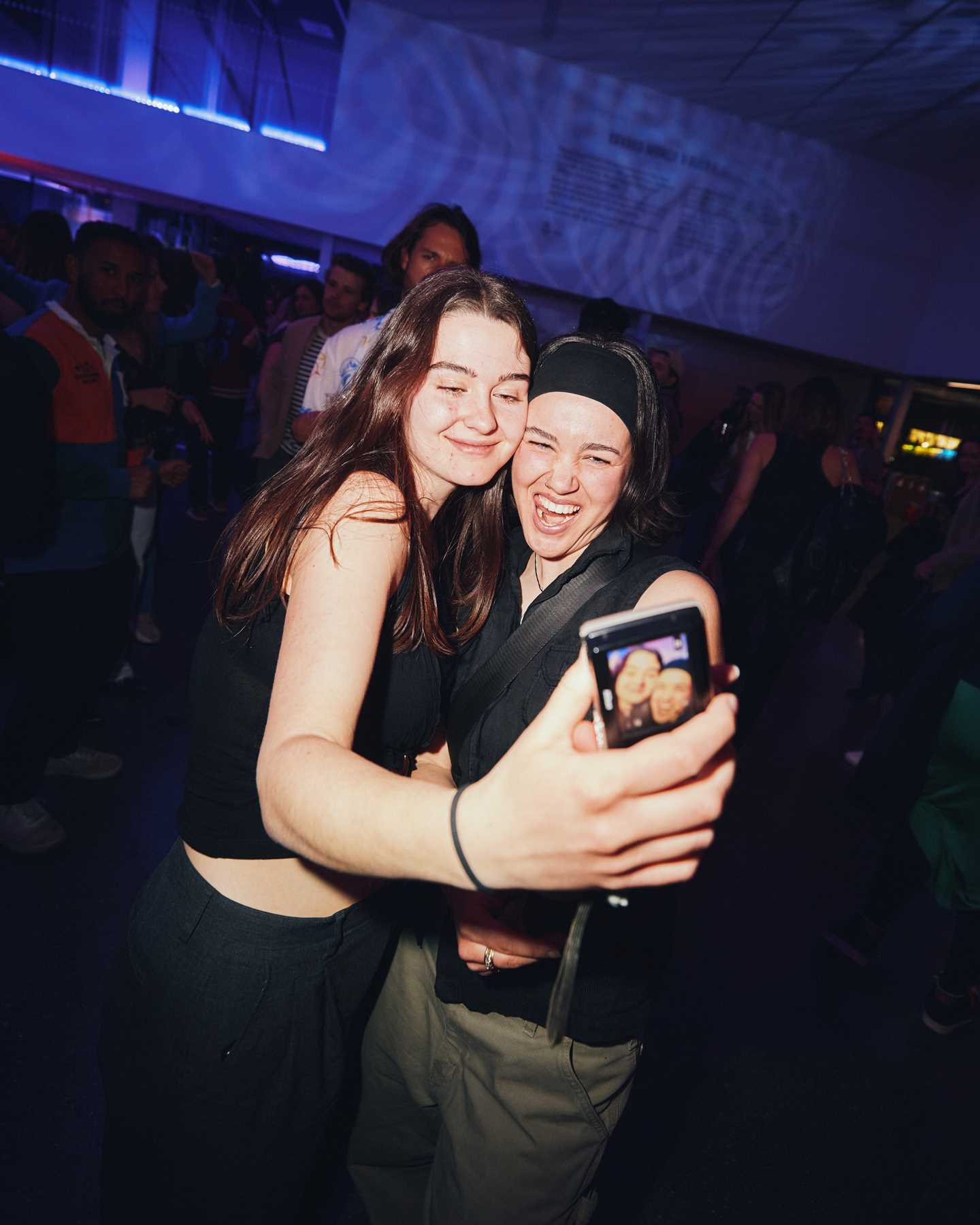 To venninner står på dansegulvet og holder rundt hverandre mens den ene av dem tar et bilde med telefonen. Begge ser inn i telefonkameraet. Fotografi