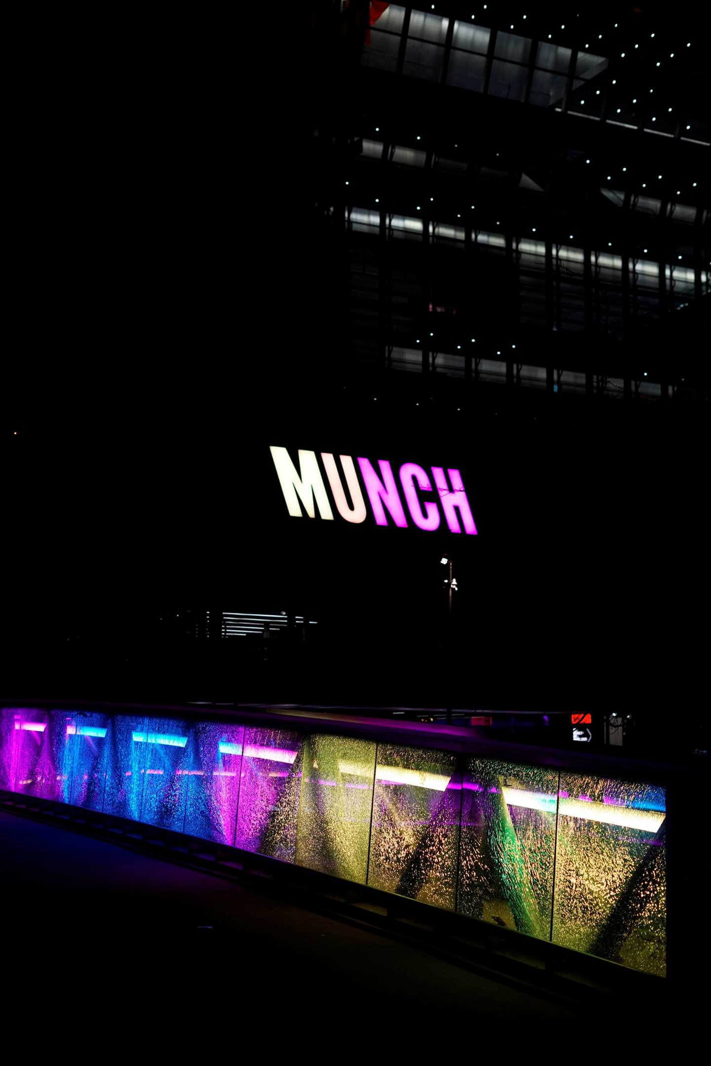 Fasaden av MUNCH-bygget i mørket. Logoen lyser i gult og rosa og det samme gjør broen foran bygget. Fotografi