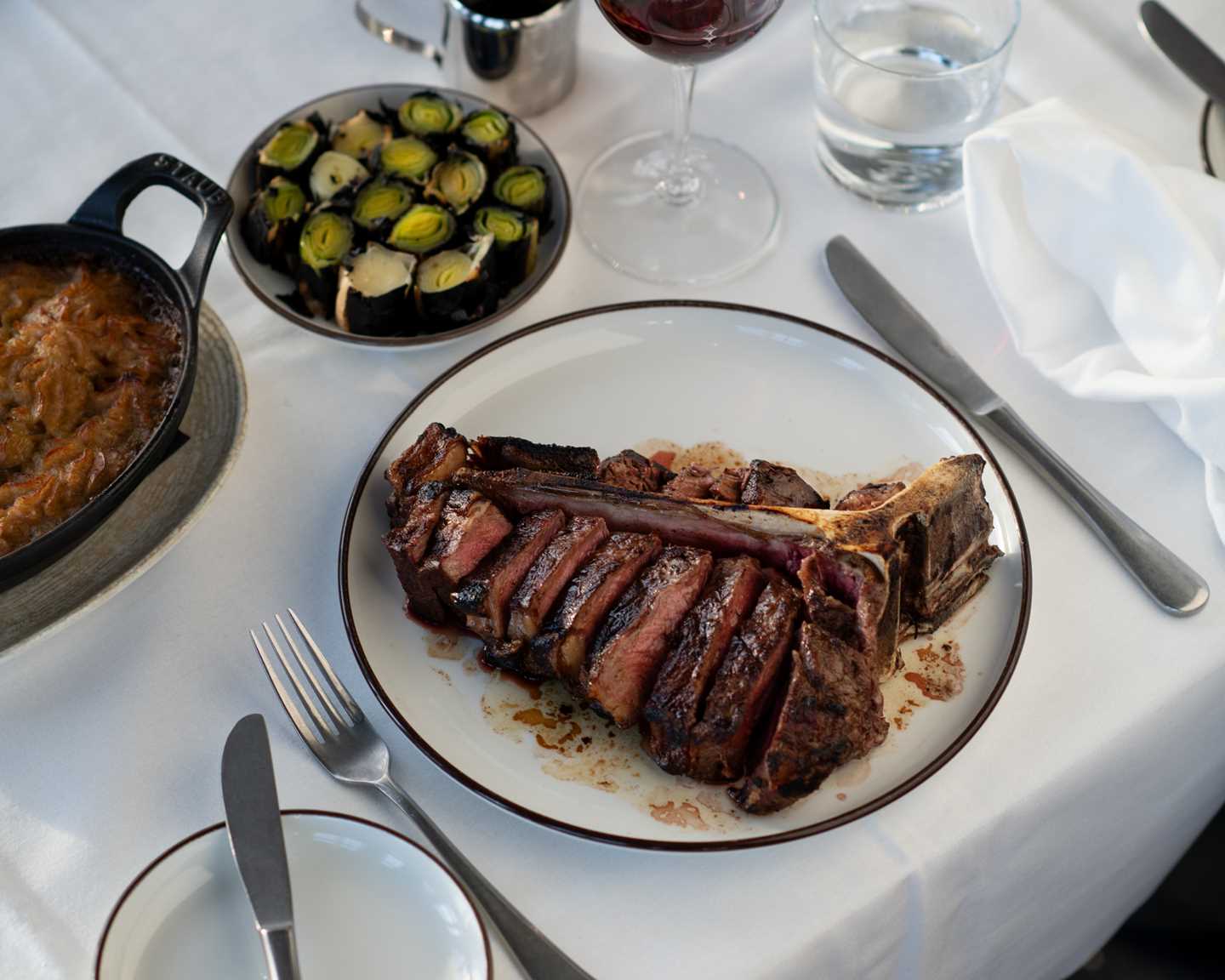 Et bord dekket med hvit duk, bestikk og stettglass. I fokus er en tallerken med en kraftig t-bone-steak. Ved siden av står et vinglass med rødvin, en tallerken med bakte purrebiter og en ildfast form poteter. Fotografi