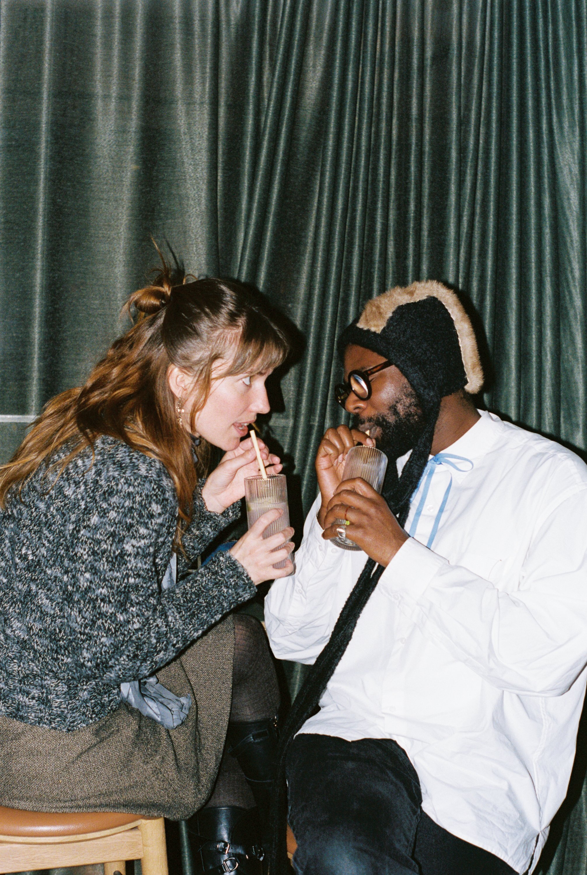 En kvinne og mann sitter tett, vendt mot hverandre og drikker av hver sin drink med sugerør mens kvinnen stirrer intenst på mannen, som har lukkede øyne. Fotografi 