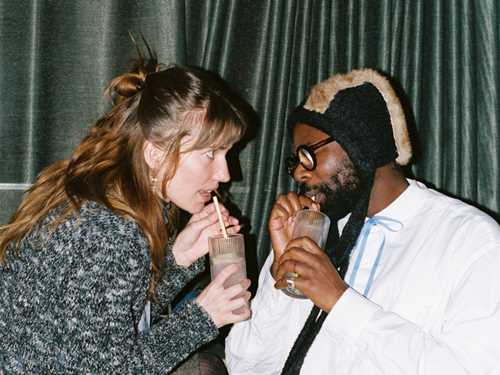 En kvinne og mann sitter tett, vendt mot hverandre og drikker av hver sin drink med sugerør mens kvinnen stirrer intenst på mannen, som har lukkede øyne. Fotografi