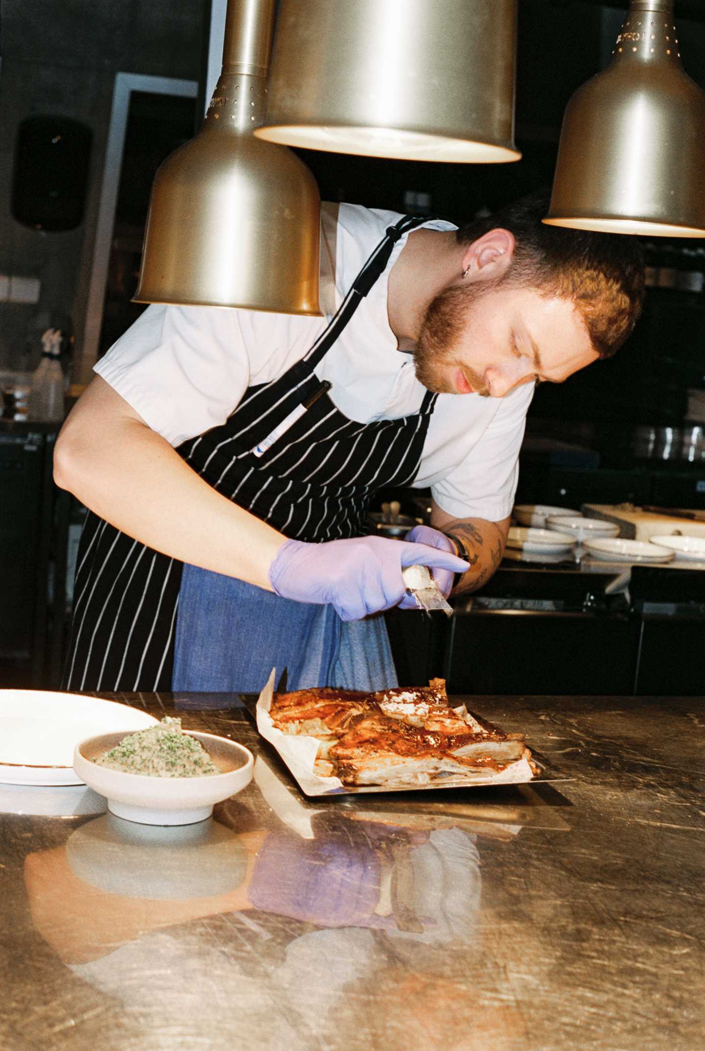 En kokk står krumbøyd over en rett mens han river ost over den. Ned fra taket over og foran kokken henger varmelamper. Fotografi