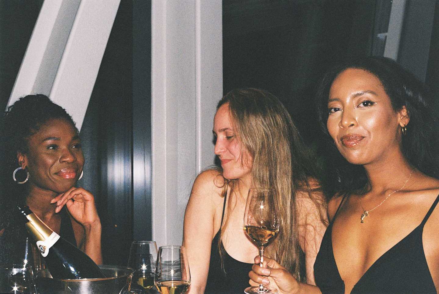 Tre kvinner sitter og står rundt et bord med flasker og glass på. Kvinnen til høyre i bildet ser rett i kamera, mens kvinnen i midten ser ned mot gulvet og kvinnen til venstre i bildet ser på kvinnen som ser inn i kameraet. Fotografi