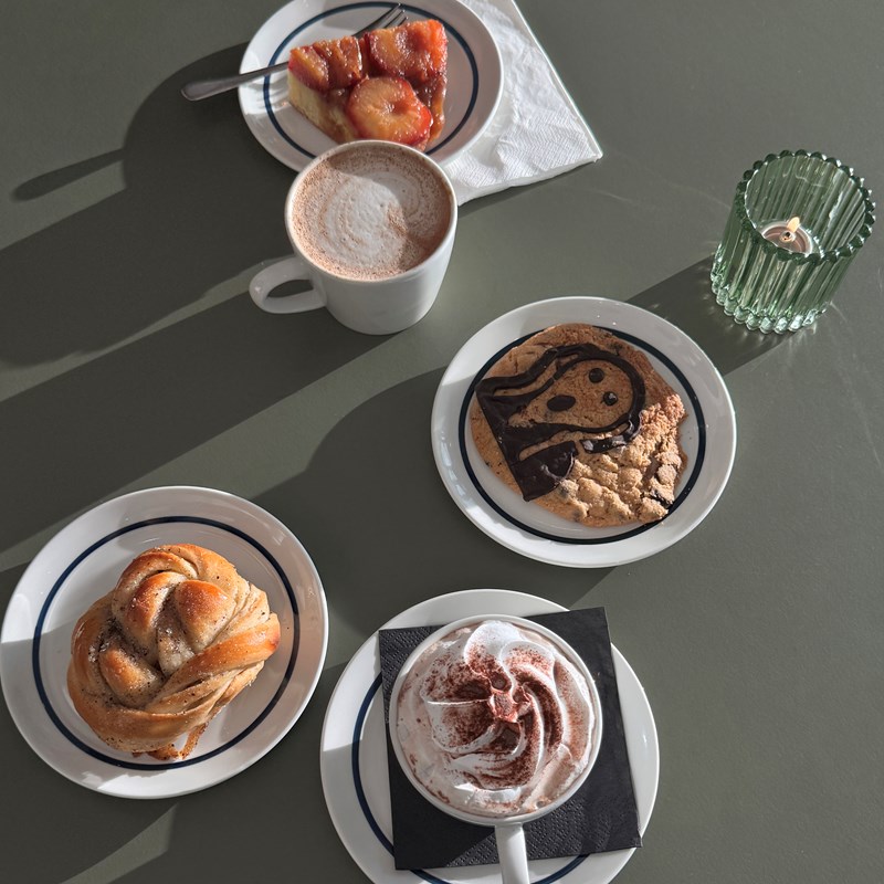 A green table seen from above, with a cup of mocha, a hot chocolate with cream, and three plates holding a cardamom bun, a cookie with The Scream motif, and a plum tart. Photograph.