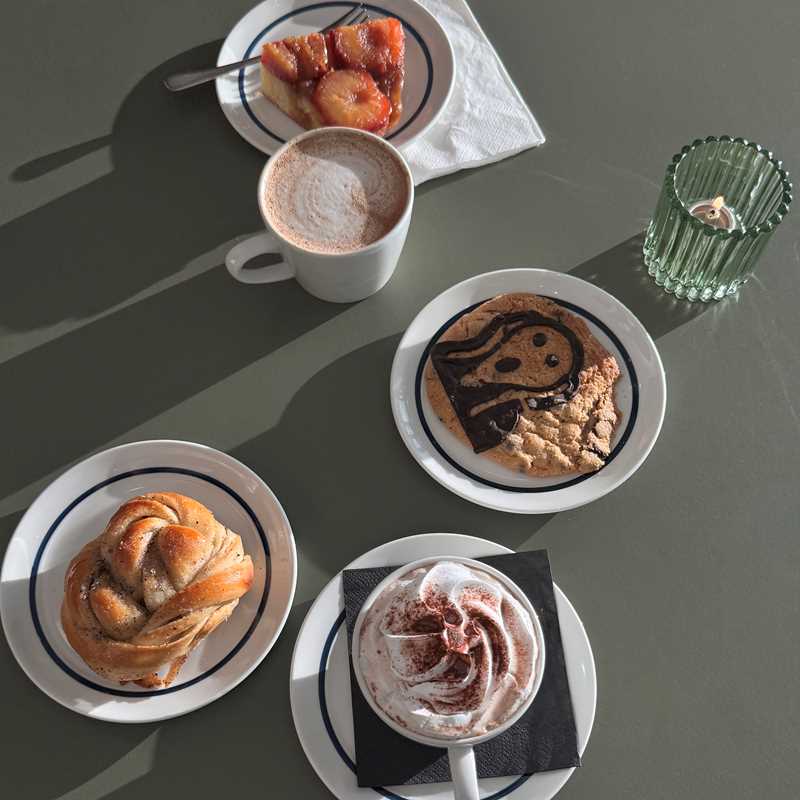 Et grønt bord sett ovenfra hvor det står en kopp mokka og en varm sjokolade med krem og tre tallerkener med kardemommebolle, cookie med Skrik-motiv og plommeterte. Fotografi
