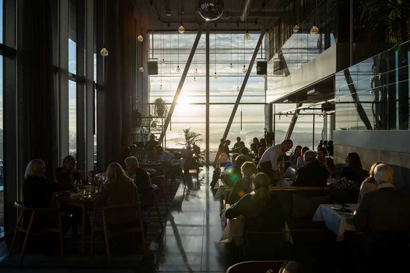 MUNCH restaurant bathed in sunlight streaming through the wall of windows at the end of the room. The restaurant is full, with people at every table and waiters moving between them. Photograph.
