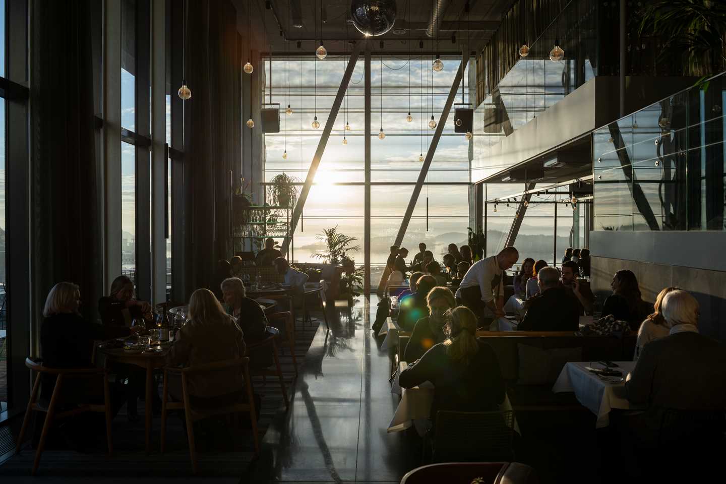 MUNCH restaurant badet i solskinn som kommer inn gjennom veggen av vinduer i enden av lokalet. Vi ser en full restaurant med mennesker ved alle bord og servitører som går rundt mellom bordene. Fotografi