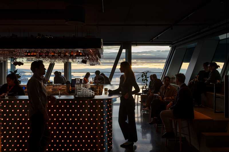 MUNCH’s bar at the top of the building. A woman and a man stand at opposite ends of the bar, while various groups of people sit around. Outside the windows, the sunset is visible, with a glimpse of the Oslo Fjord. Photograph.