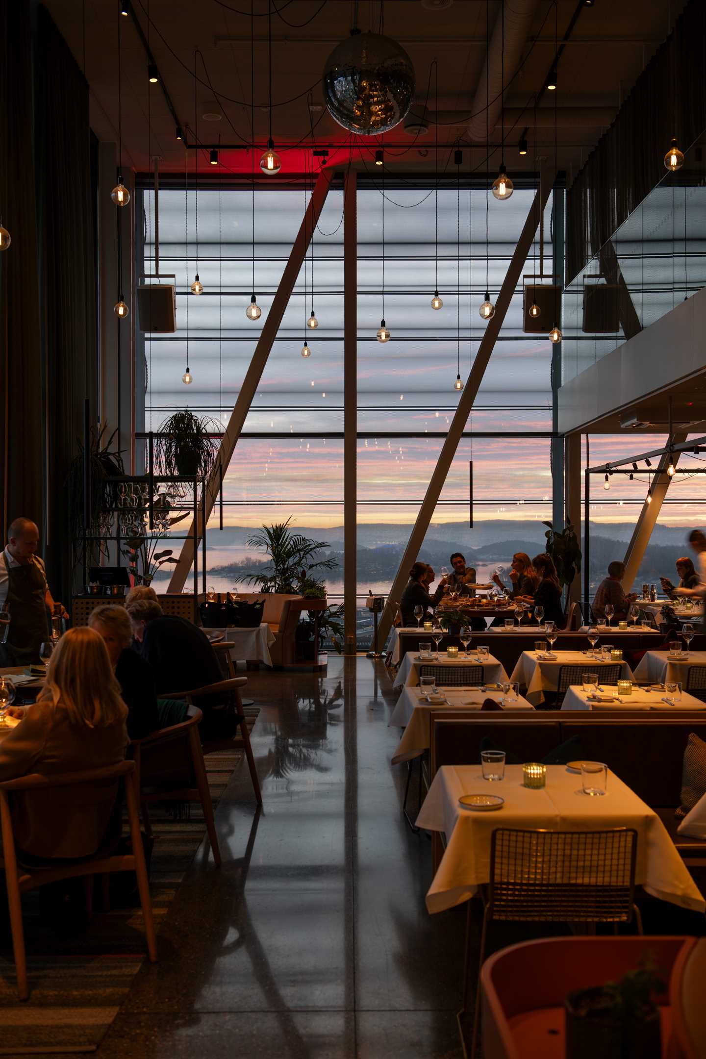 A half-full restaurant with small and large tables covered in white tablecloths, and several groups seated at different tables. At the far end, floor-to-ceiling windows reveal the sunset and the Oslo Fjord outside. Photograph.