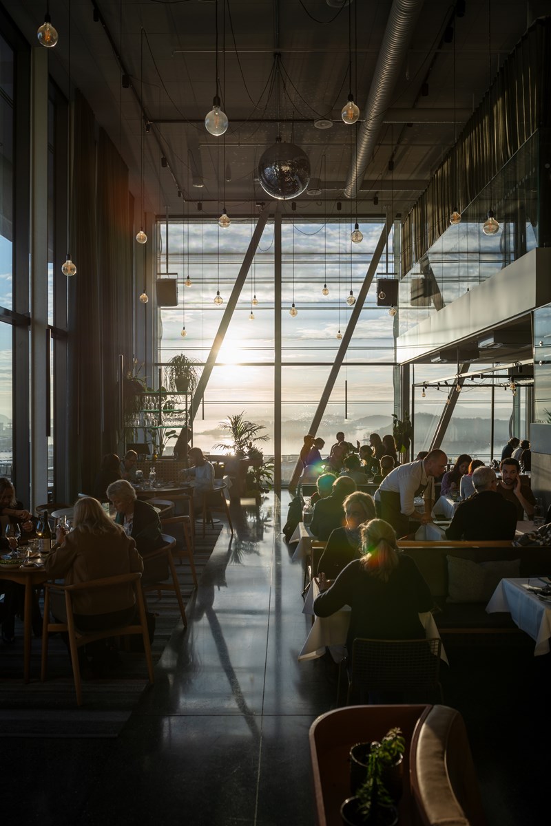 MUNCHs restaurantlokale i byggets øverste etasje. Vi ser en full restaurant med mennesker ved alle bord og kelnere som snakker med noen av gjestene. I enden av lokalet er det vinduer fra gulv til tak som solen skinner inn gjennom og vi ser utsikten utover Oslofjorden. Fotografi