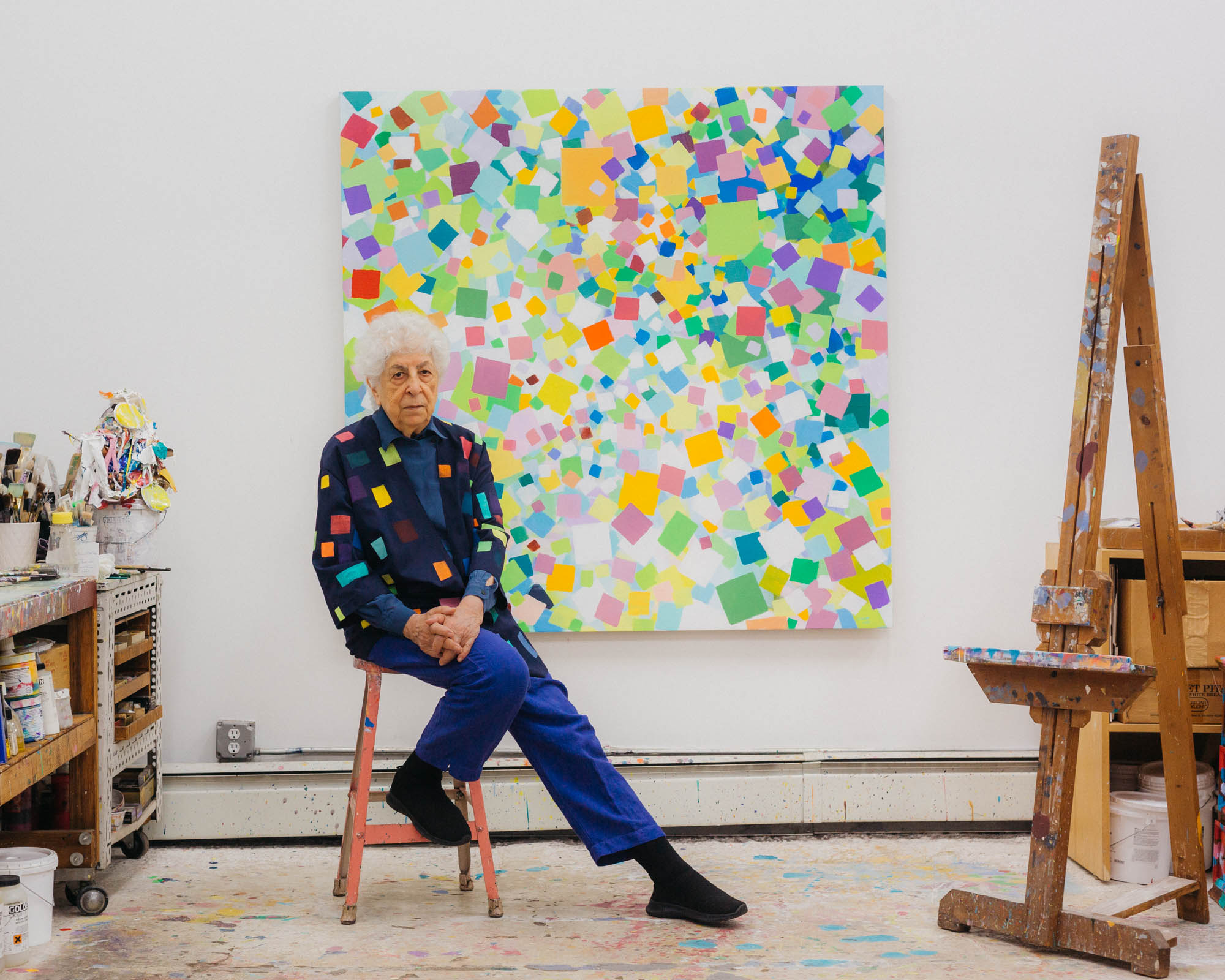 The artist Samia Halaby sits on a pink stool in her studio in front of one of her own works. The wall is white and the artwork consists of many colourful squares. To the right of the image stands an easel with paint stains, and to the left we see a shelving unit full of paint and painting materials. Photograph.
