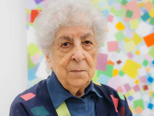 Portrett av kunstneren fra livet og opp, stående foran et av sine egne kunstverk. Fotografi