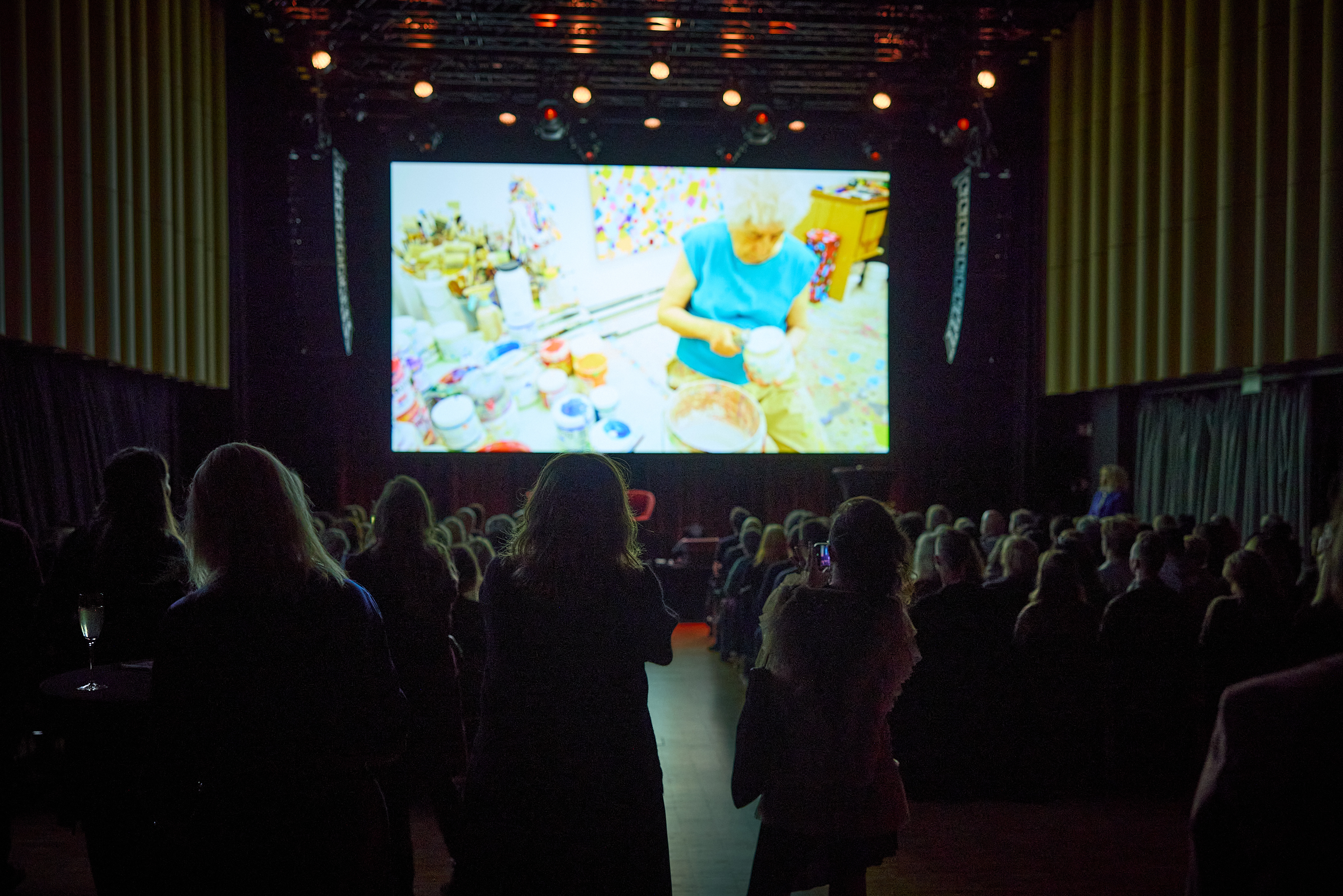 A packed banquet hall seen from the opposite side of the stage. All the rows of chairs are filled, and many people are standing in front of the camera. Everyone is watching a large screen behind the stage showing a film. Photograph.