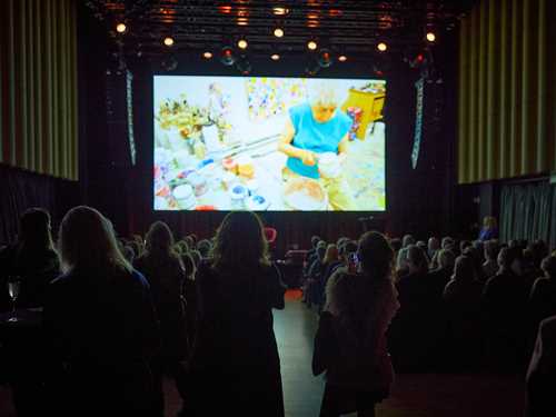 A packed banquet hall seen from the opposite side of the stage. All the rows of chairs are filled, and many people are standing in front of the camera. Everyone is watching a large screen behind the stage showing a film. Photograph.
