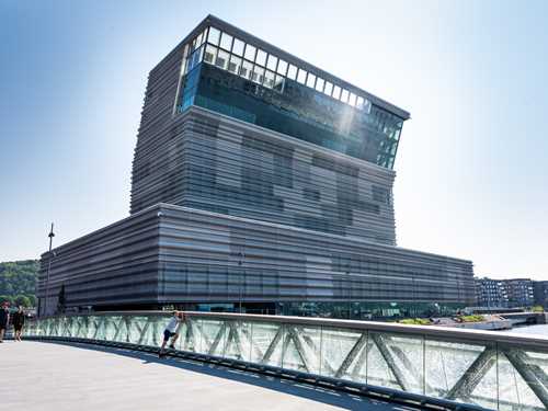 The new MUNCH in Bjørvika, seen from the west in a low-angle view. The sun is shining, and the sky is blue. In the foreground, you can see the bridge crossing the Akerselva River in front of the museum. Photograph.