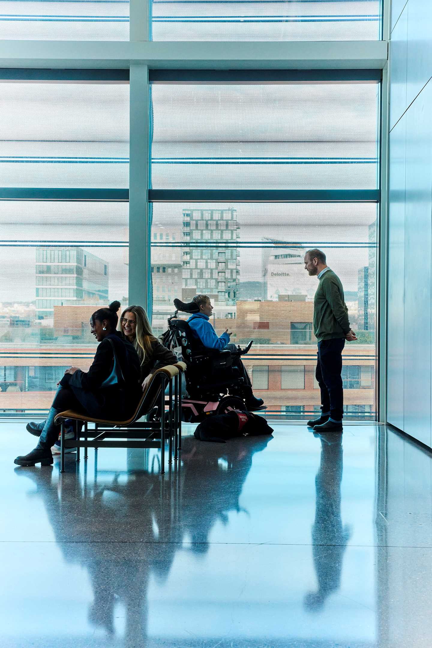 Two women sit laughing on a bench outside an exhibition room at MUNCH. Behind them, a woman in a wheelchair talks with a man. On the floor beside the wheelchair lies a service dog. Photograph.
