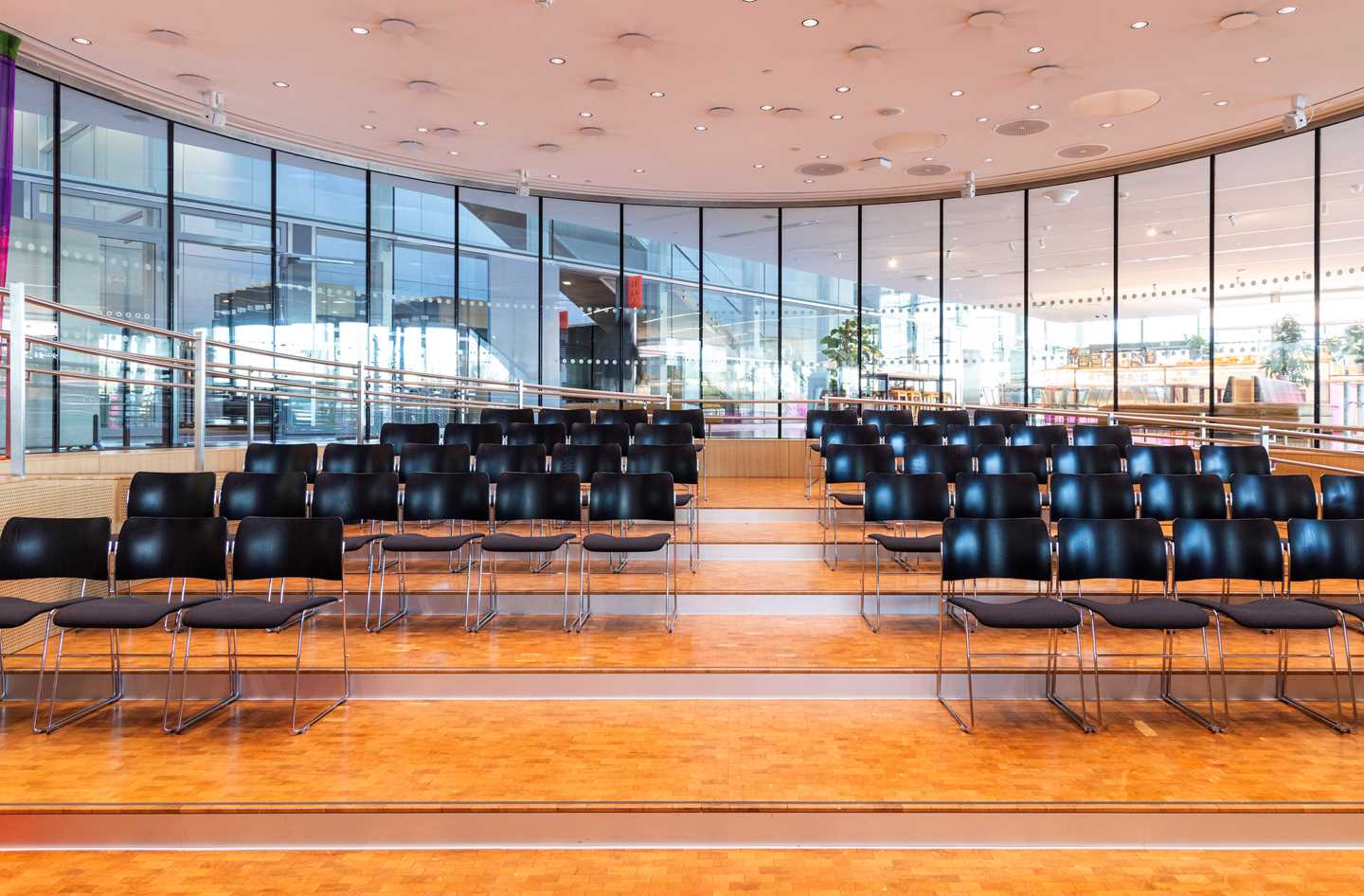 The amphitheatre at MUNCH seen from the podium at the bottom. Four rows of black chairs are separated by a central aisle. Windows surround the entire room. Photograph.