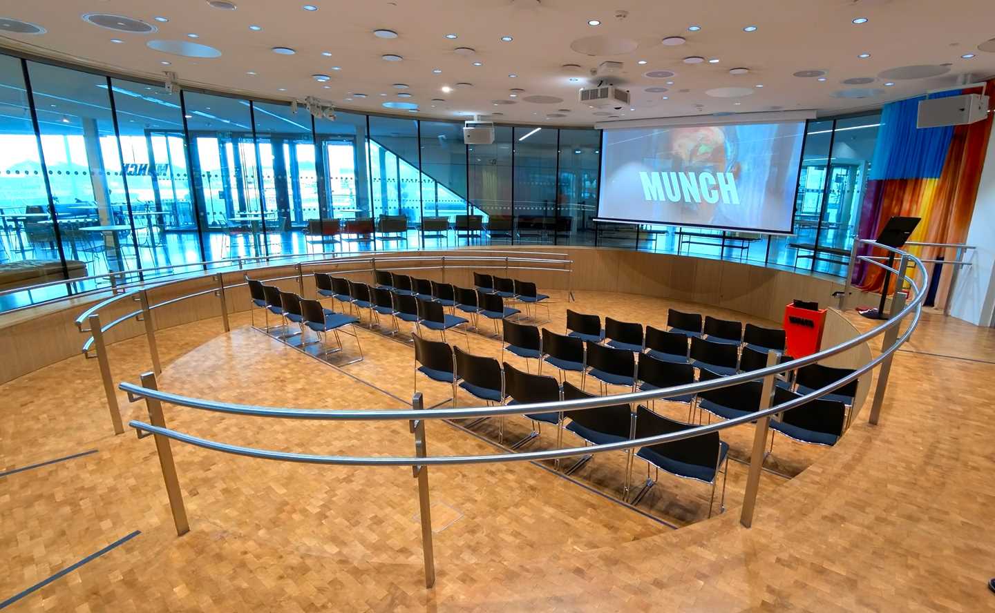 The Amfi hall at MUNCH. A circular room with windows all around. In the middle are four rows of chairs facing a screen hanging from the ceiling. Photograph.