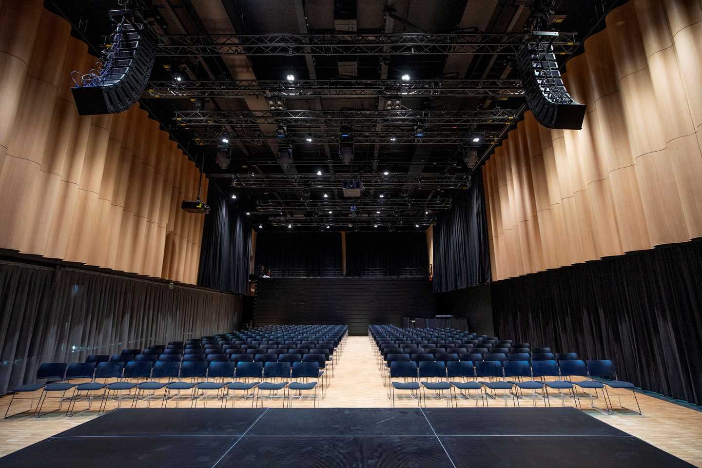 The banquet hall at MUNCH seen from the stage. A large sound and lighting rig hangs from the ceiling. On the floor are many rows of black chairs facing the stage. Photograph.