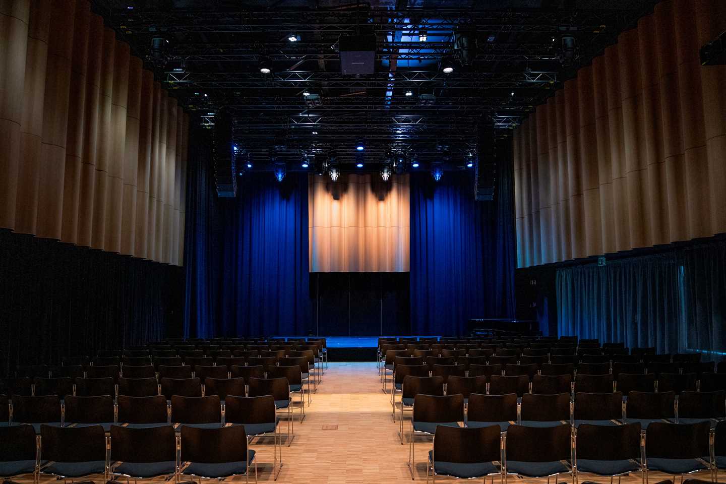 The banquet hall at MUNCH seen from the back. Several rows of black chairs face a stage illuminated with blue stage lighting. Photograph.