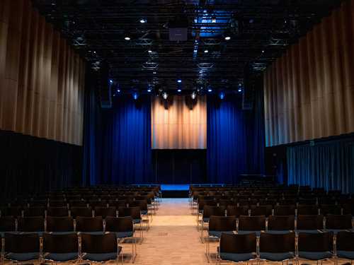 Festsalen på MUNCH sett fra bakerst i lokalet. Det er flere stolrader med svarte stoler som står foran en scene med blått scenelys. Fotografi