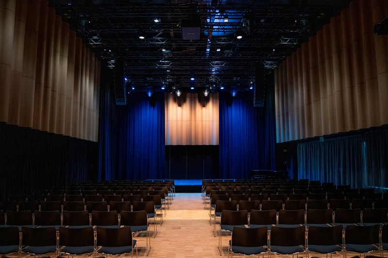 The banquet hall at MUNCH seen from the back. Several rows of black chairs face a stage illuminated with blue stage lighting. Photograph.