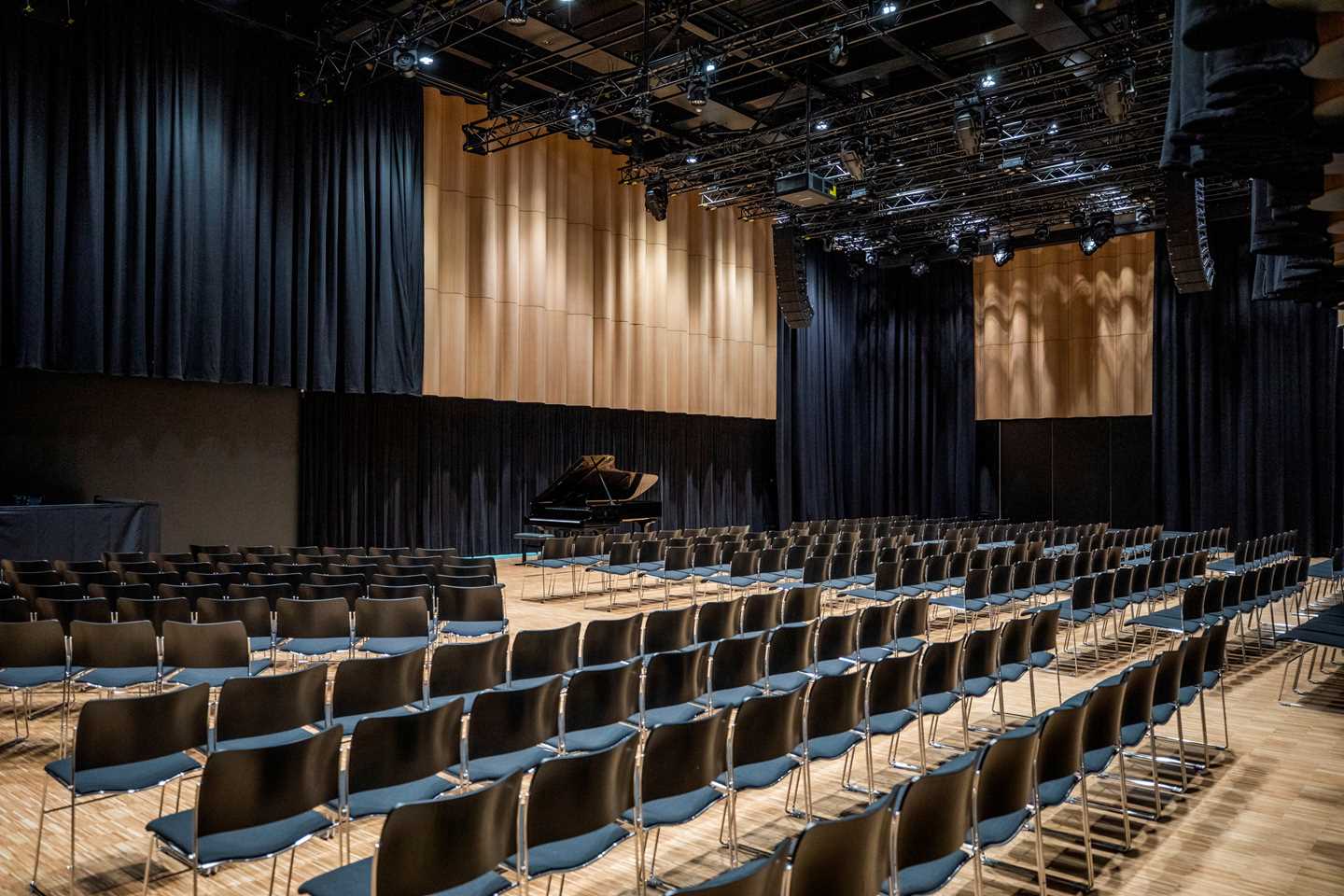 The banquet hall at MUNCH seen from the right side. Several rows of black chairs face a grand piano positioned along one of the hall’s long walls. Photograph.