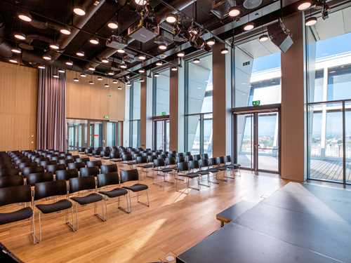 The top hall at MUNCH seen from the stage. Inside the room are rows of black chairs, and one wall consists of floor-to-ceiling windows, with a rooftop terrace visible outside. Photograph.